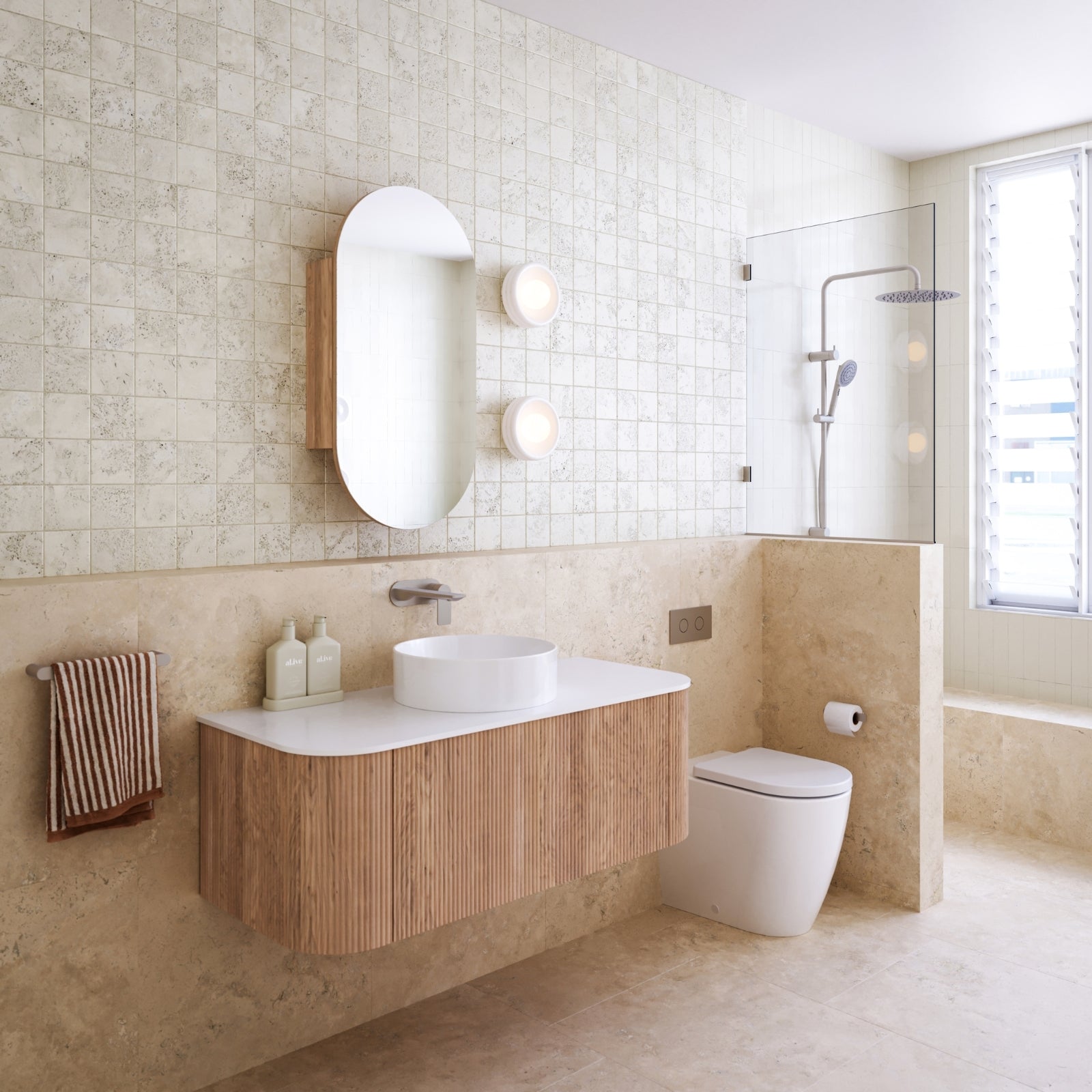 Coastal style bathroom with wooden vanity, white sink, and toilet against a travertine tiled wall.