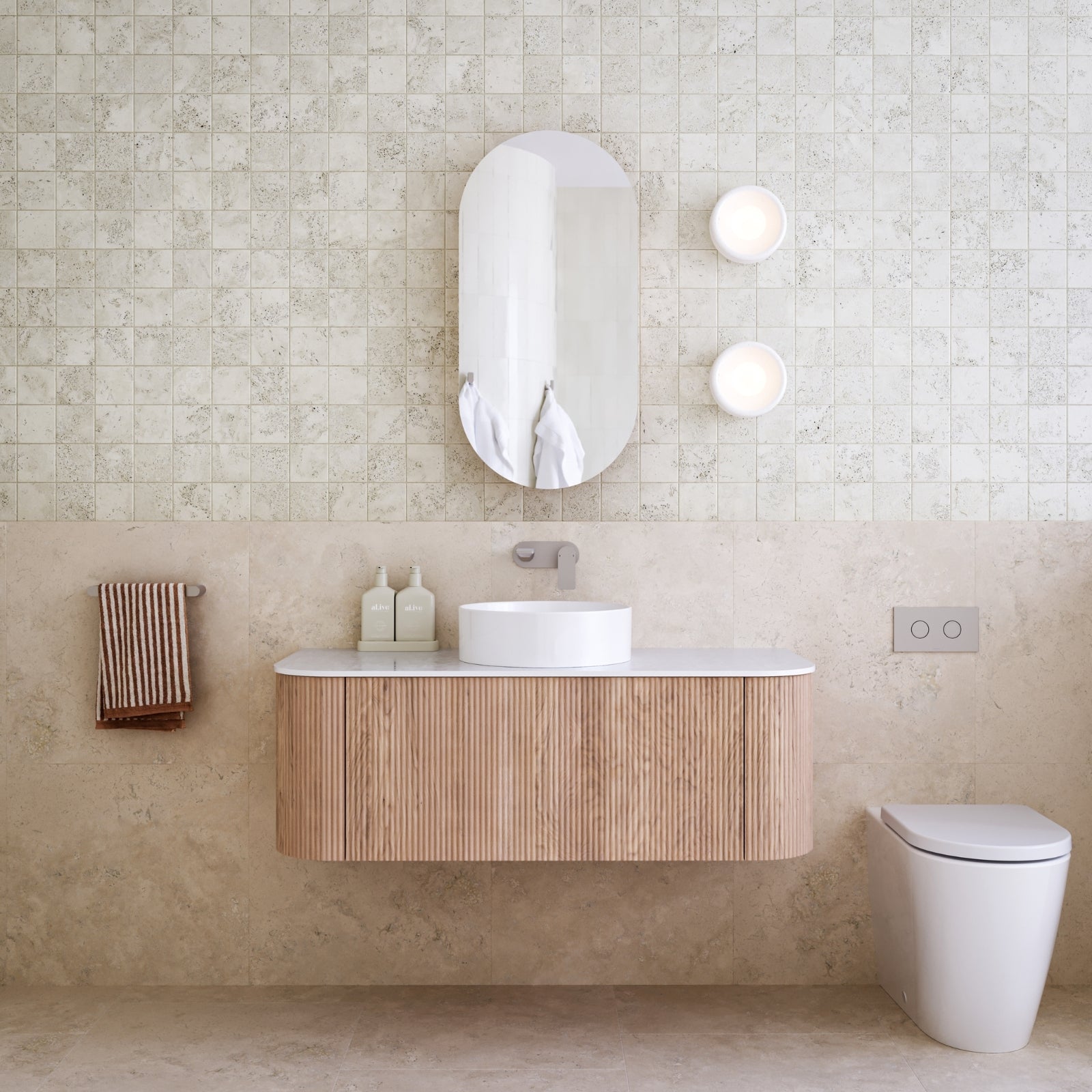 Coastal style bathroom with wooden vanity, white sink, and toilet against a travertine tiled wall.